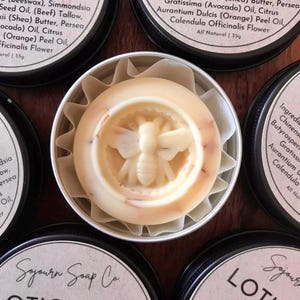 May include: A round, white lotion bar with a bee design in the centre, packaged in a silver tin. The label on the tin reads "Sojourn Soap Co. Lotion Bar Orange" and lists the ingredients.