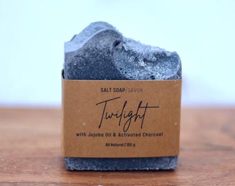 May include: A bar of charcoal soap with a brown paper label that reads "SALT SOAP/SAVON Twilight with Jojoba Oil & Activated Charcoal All Natural | 180 g".