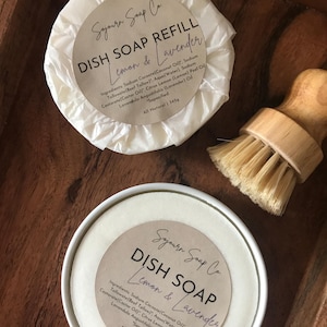 May include: Two white containers of dish soap with a brown wooden brush. The containers are labelled "Sojourn Soap Co. Dish Soap Refill Lemon & Lavender" and "Sojourn Soap Co. Dish Soap Lemon & Lavender". The labels list the ingredients and state that the soap is all natural and 345g.