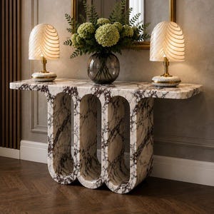 May include: A white and brown marble console table with three arched cutouts, topped with two glass lamps featuring gold-toned ribbed shades and a clear glass vase filled with green hydrangea flowers and leafy foliage.