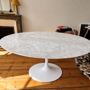 May include: An oval dining table featuring a white marble top with grey veining and a smooth, white, pedestal-style base. The table surface is polished and reflective, showcasing the natural stone pattern across the entire tabletop area.