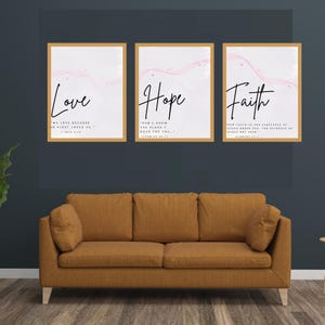 May include: Three framed wall art prints with the words "Love", "Hope", and "Faith" in elegant script. Each print has a gold frame and a pink watercolor design. The prints are displayed above a brown sofa.