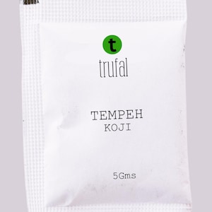 May include: A white packet of Trufal tempeh koji with a green logo. The text "TEMPEH KOJI" is printed in black, along with "5 Gms". The packet has a textured surface and a dark brown and green sealed edge.