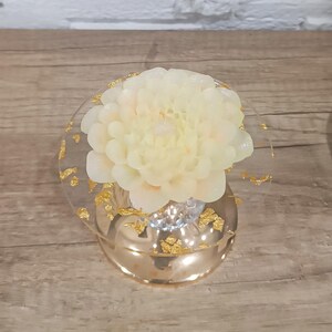 Puede incluir: Una vela de flor blanca sobre un soporte de vidrio dorado y transparente. El soporte tiene una parte superior redonda y una base con detalles de pan de oro.