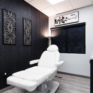 May include: A white medical examination table with adjustable height and footrest in a room with black walls and gold accents. The room has a window with black blinds, a black cabinet, and a gold metal shelf with plants and decorative items.