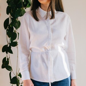 Op de afbeelding: Een wit overhemd met lange mouwen en een strik in de taille, gedragen met een blauwe jeans. Het overhemd heeft een klassieke kraag en een knoopsluiting. Een groene plant is zichtbaar op de achtergrond. De stof is licht.