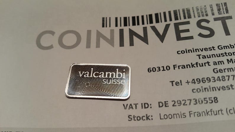Puede incluir: Una peque&ntilde;a barra rectangular de plata con el logotipo "valcambi suisse", sobre un documento con el logotipo "COININVEST" y otro texto. La barra tiene un dise&ntilde;o geom&eacute;trico.