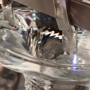 Puede incluir: Primer plano de un l&iacute;quido transparente que gira en un vaso de precipitados de vidrio. Una pinza met&aacute;lica sujeta una peque&ntilde;a pieza met&aacute;lica con la etiqueta "valcom" sobre el l&iacute;quido que gira. El vaso tiene una marca de 1500 ml.