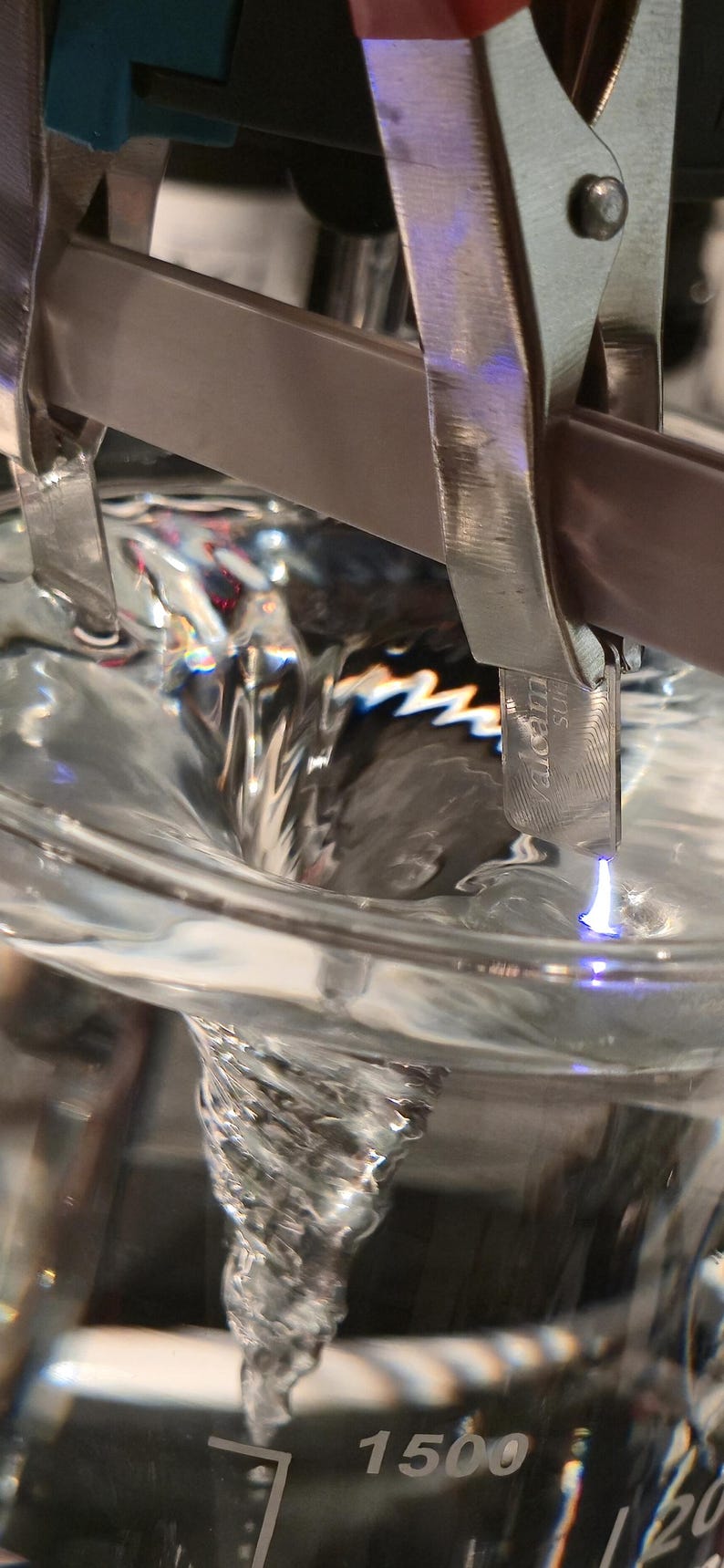 Puede incluir: Primer plano de un l&iacute;quido transparente que gira en un vaso de precipitados de vidrio. Una pinza met&aacute;lica sujeta una peque&ntilde;a pieza met&aacute;lica con la etiqueta "valcom" sobre el l&iacute;quido que gira. El vaso tiene una marca de 1500 ml.