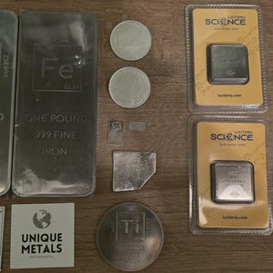 Puede incluir: Una variedad de barras, monedas y lingotes de metal, incluyendo barras de zinc y hierro de una libra. Tambi&eacute;n se muestran peque&ntilde;os cuadrados de metal, una moneda de titanio y una balanza digital que muestra 39,16. Los objetos est&aacute;n sobre una superficie de madera.