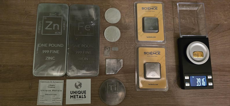 Puede incluir: Una variedad de barras, monedas y lingotes de metal, incluyendo barras de zinc y hierro de una libra. Tambi&eacute;n se muestran peque&ntilde;os cuadrados de metal, una moneda de titanio y una balanza digital que muestra 39,16. Los objetos est&aacute;n sobre una superficie de madera.