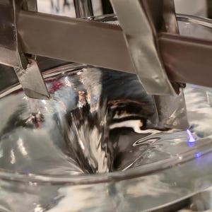 Puede incluir: Primer plano de un vaso de precipitados de vidrio transparente lleno de un l&iacute;quido arremolinado y reflectante. Unas pinzas met&aacute;licas sujetan una barra marr&oacute;n y blanca sobre el l&iacute;quido. La imagen destaca la textura del l&iacute;quido y el entorno de laboratorio.