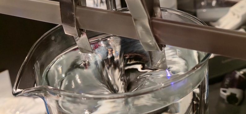 Puede incluir: Primer plano de un vaso de precipitados de vidrio transparente lleno de un l&iacute;quido arremolinado y reflectante. Unas pinzas met&aacute;licas sujetan una barra marr&oacute;n y blanca sobre el l&iacute;quido. La imagen destaca la textura del l&iacute;quido y el entorno de laboratorio.