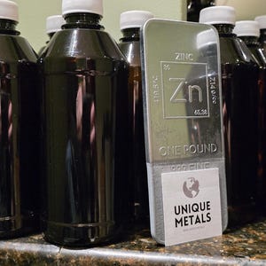 Puede incluir: Varias botellas marrones oscuras con tapas blancas junto a una barra de zinc plateada. La barra est&aacute; marcada con los detalles del elemento, el peso y el texto "ONE POUND". Una etiqueta dice "UNIQUE METALS".