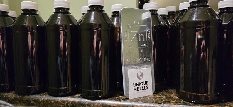 Puede incluir: Varias botellas marrones oscuras con tapas blancas junto a una barra de zinc plateada. La barra est&aacute; marcada con los detalles del elemento, el peso y el texto "ONE POUND". Una etiqueta dice "UNIQUE METALS".