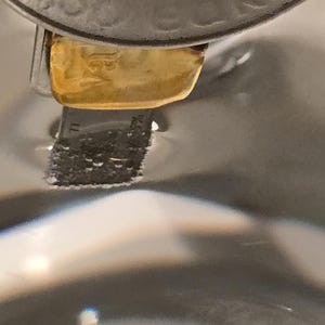 Puede incluir: Primer plano de un objeto de metal plateado con una pieza rectangular dorada adjunta. El objeto parece estar parcialmente sumergido en un l&iacute;quido transparente, con un fondo borroso.