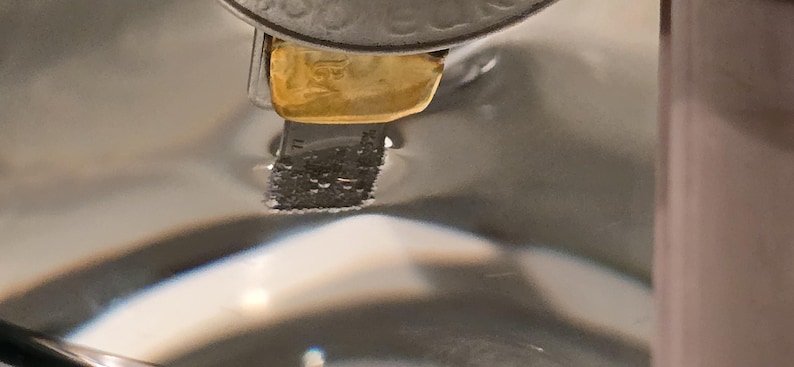 Puede incluir: Primer plano de un objeto de metal plateado con una pieza rectangular dorada adjunta. El objeto parece estar parcialmente sumergido en un l&iacute;quido transparente, con un fondo borroso.
