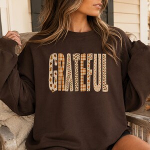 May include: A dark brown crewneck sweatshirt featuring the word GRATEFUL printed across the chest. Each letter is filled with a different pattern, including acorns, orange plaid, green leaves, leopard print, and small orange floral designs.