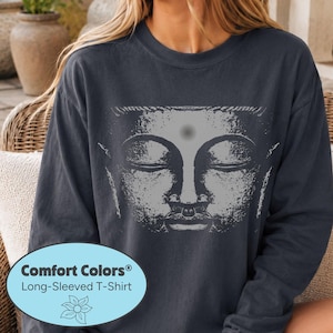 May include: A dark charcoal gray long-sleeved t-shirt featuring a large, light gray graphic print of a serene Buddha face with closed eyes and a central dot on the forehead. The shirt has a crew neckline and a relaxed, casual fit.