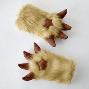 May include: A pair of plush, furry gloves designed to resemble animal paws. The gloves are primarily tan with brown accents on the claws and a star-shaped detail on the palm. The gloves are made of soft, faux fur material.