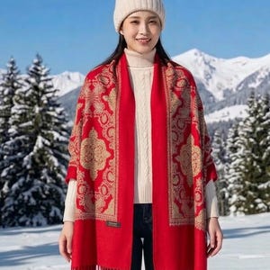 May include: A red scarf with a gold floral pattern and fringe, worn with a cream-coloured beanie and sweater. The scarf is draped over the shoulders. The background features snow-covered trees and mountains.