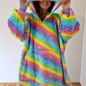 Può includere: Una grande coperta con cappuccio con un motivo a arcobaleno e stelle bianche e una grande tasca sul davanti.
