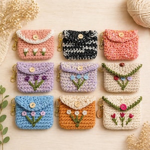 May include: Nine handmade crochet coin purses in various colors, including pink, black, purple, blue, and orange. Each purse features a button closure, a floral design, and a gold-colored keychain. The purses are displayed on a light wooden surface.
