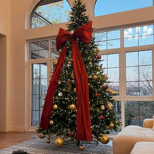 Può includere: Un albero di Natale decorato con un grande fiocco rosso e ornamenti dorati. L'albero è in una stanza luminosa con una grande finestra. Un cane nero è sdraiato su un tappeto fantasia davanti all'albero.