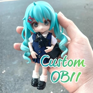 May include: A custom OB11 doll with turquoise pigtails, wearing a navy blue dress, white shirt, and black shoes. The doll has a black satchel and red hair clips. The text "Custom OB11" is overlaid on the image.