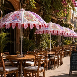 May include: A series of outdoor patio umbrellas featuring white fabric with intricate, colorful floral and bird patterns, scalloped red and white trim, and white poles, positioned over wooden tables and chairs at a sidewalk cafe.