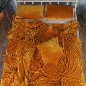 Puede incluir: Una cama con estructura metálica blanca está cubierta con un juego de cama de terciopelo amarillo mostaza. El juego incluye una funda nórdica y tres fundas de almohada a juego. La ropa de cama tiene un aspecto suave y lujoso.
