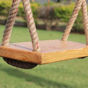 May include: A wooden swing set with natural wood grain and two thick, knotted ropes. The ropes are tied to the top of the swing set and hang down to the wooden seat. The seat is rectangular and has a slightly rounded edge.