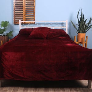 Op de afbeelding: Een bed met een wit metalen frame is bedekt met een dieprode, zachte deken en bijpassende kussens. De kamer heeft een blauwe muur, houten luiken en potplanten, wat een gezellige slaapkameromgeving creëert.
