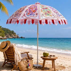 May include: A beach umbrella featuring a white canopy with colorful embroidered peacock motifs and red scalloped trim. The umbrella has a central pole and is positioned over a wooden chair, a straw bag, and a small wooden table with a coconut.