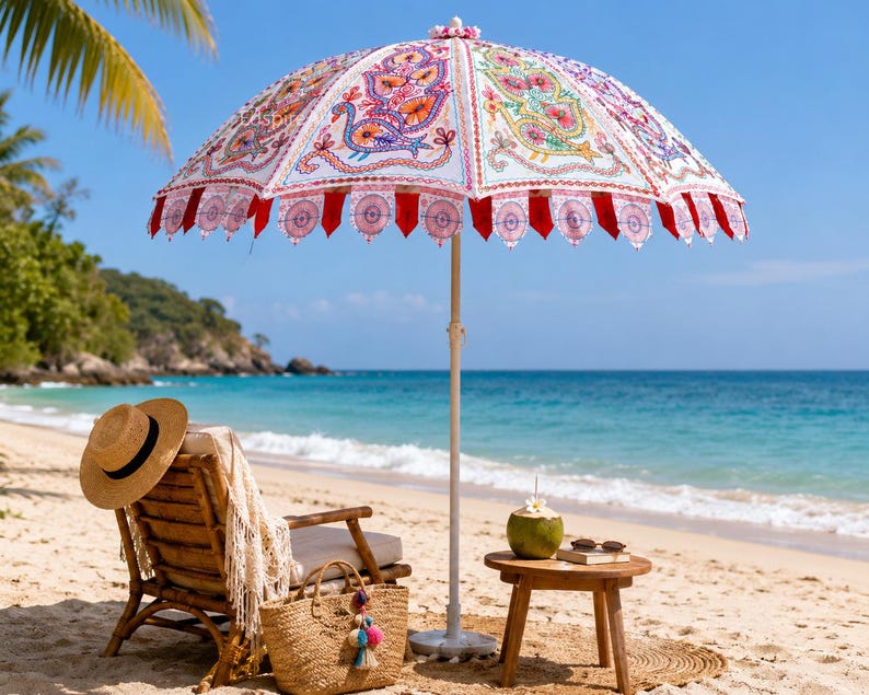May include: A beach umbrella featuring a white canopy with colorful embroidered peacock motifs and red scalloped trim. The umbrella has a central pole and is positioned over a wooden chair, a straw bag, and a small wooden table with a coconut.