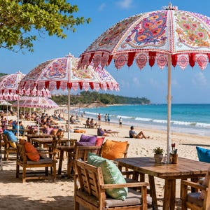May include: A series of large beach umbrellas with white fabric featuring colorful, intricate paisley patterns and red scalloped trim. The umbrellas are positioned over wooden tables and chairs with various colored cushions on a sandy beach.