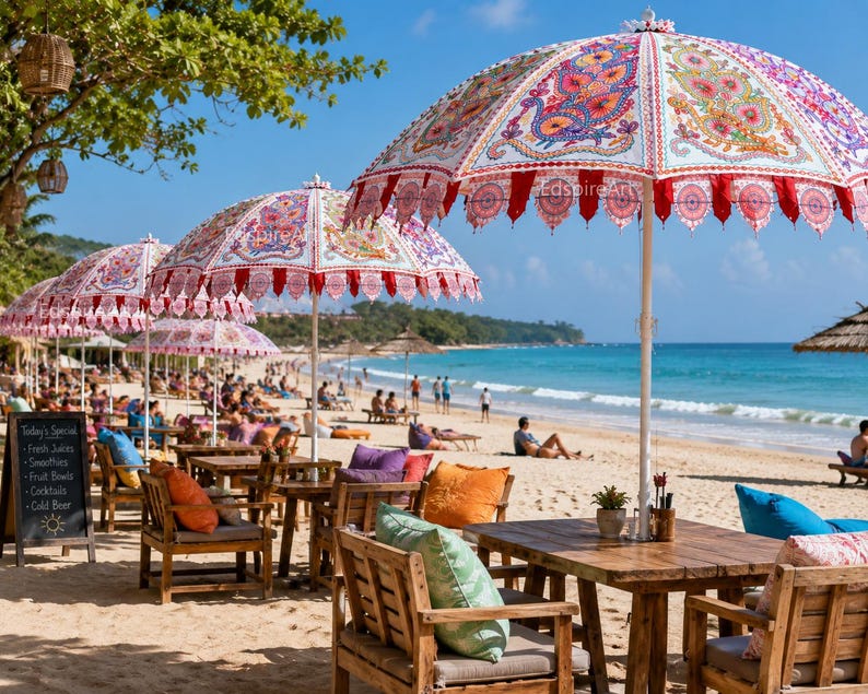 May include: A series of large beach umbrellas with white fabric featuring colorful, intricate paisley patterns and red scalloped trim. The umbrellas are positioned over wooden tables and chairs with various colored cushions on a sandy beach.