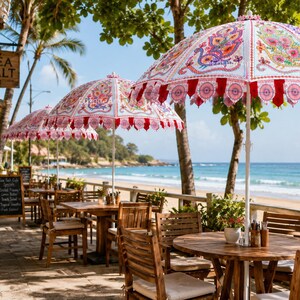 May include: A series of white patio umbrellas featuring intricate, colorful peacock-inspired patterns in shades of pink, orange, and blue, with red scalloped trim, positioned over wooden tables and chairs on a stone terrace overlooking the ocean.