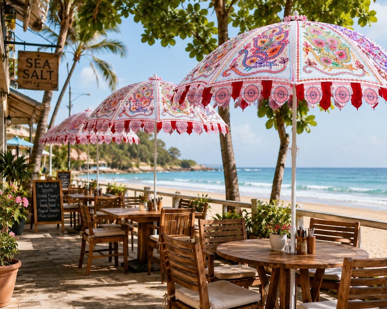 May include: A series of white patio umbrellas featuring intricate, colorful peacock-inspired patterns in shades of pink, orange, and blue, with red scalloped trim, positioned over wooden tables and chairs on a stone terrace overlooking the ocean.