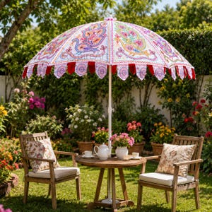 May include: A large white garden umbrella featuring colorful embroidered peacock motifs and decorative scalloped edges with red fabric accents. The umbrella is positioned over a round wooden table with a white teapot and two chairs with floral pillows.