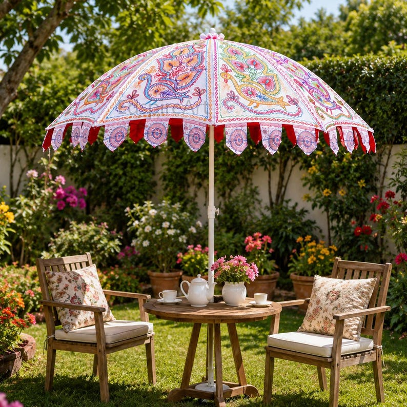 May include: A large white garden umbrella featuring colorful embroidered peacock motifs and decorative scalloped edges with red fabric accents. The umbrella is positioned over a round wooden table with a white teapot and two chairs with floral pillows.