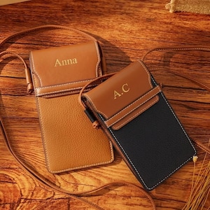Puede incluir: Dos fundas verticales para teléfono tipo bandolera con correas largas y finas. Una funda es de color tostado con el nombre Anna grabado en la solapa. La segunda es negra con solapa tostada y las iniciales A.C. grabadas. Costuras visibles.
