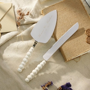 May include: A silver-toned cake server and matching serrated knife set featuring white, pearl-like beaded handles. The metal blades are engraved with the text Monroe & Matthew 12.10.2023. The utensils have a polished finish and a classic design.