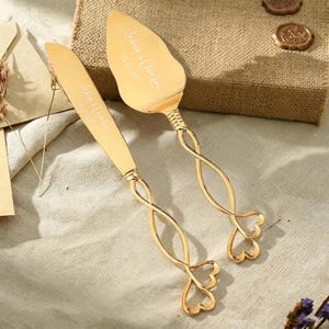 Peut inclure: Un ensemble de couteau et pelle à gâteau doré avec le texte gravé Ashley & Christian 10.15.2022. Les deux ustensiles possèdent de longs manches fins au design métallique torsadé et ouvert, se terminant par deux boucles en forme de cœur.