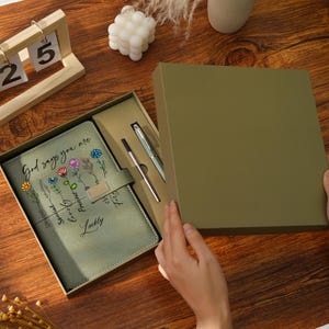May include: A sage green journal and pen set in a gift box. The journal features floral illustrations and the text "God says you are..." The set includes a silver pen and a black pen. The box is olive green.