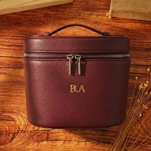 Peut inclure: Une trousse de toilette bordeaux à la texture grainée, dotée d'une poignée supérieure et d'une double fermeture éclair argentée. L'avant de la trousse présente les initiales B.A. brodées en doré au centre de la surface extérieure structurée.