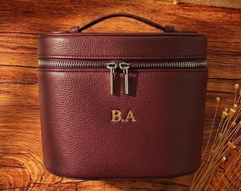 May include: A deep burgundy, pebble-textured vanity case with a top handle and a dual-zipper closure. The front of the structured, cylindrical bag features the gold-colored embossed initials B.A. centered on the exterior surface.