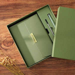 May include: A sage green gift set featuring a notebook and two pens, presented in a matching box. The notebook has a gold nameplate that reads "Patricia". The set is displayed on a wooden surface.