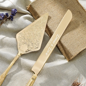 Puede incluir: Un juego de pala y cuchillo para pastel en tono dorado. La pala triangular presenta un intrincado patrón floral en relieve. El cuchillo largo y dentado tiene grabado el texto "Leonid & Lerie" y la fecha "03.15.2026" en letra cursiva.
