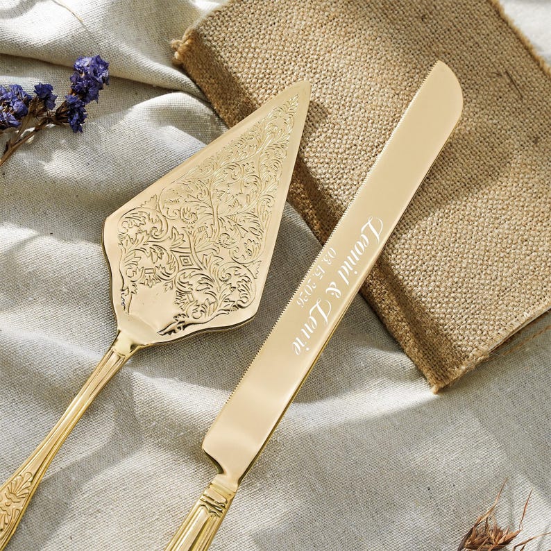 May include: A gold-toned cake server and knife set. The triangular server features an intricate embossed floral pattern. The long, serrated knife is engraved with the text "Leonid & Lerie" and the date "03.15.2026" in a cursive script font.